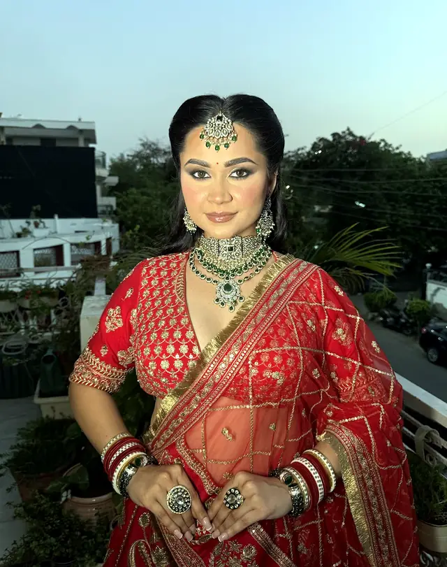 Bride in a red lehenga with a maang tikka, photographed indoors against a plain wall
