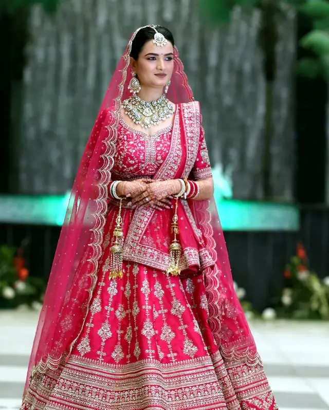 Bride in a rani-pink lehenga with a sheer dupatta drawn over her head like a veil, garden backdrop
